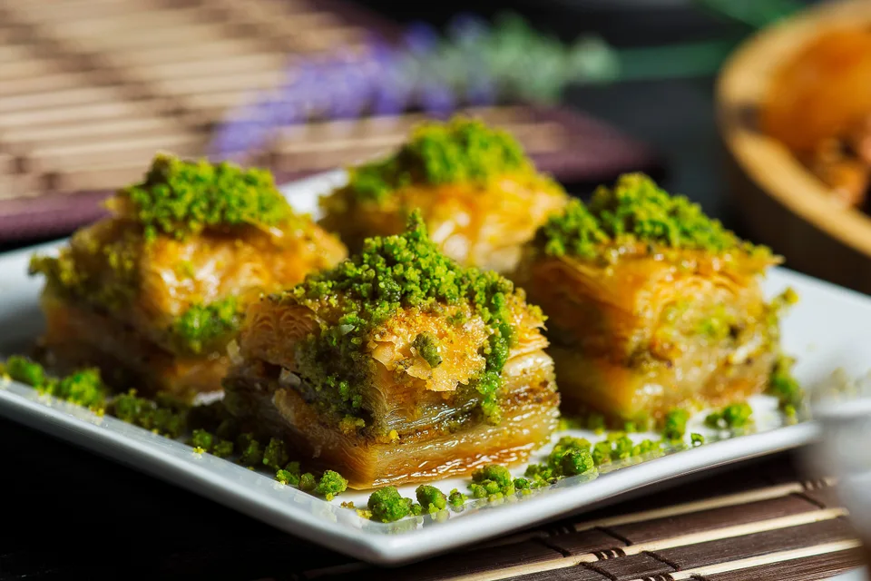 BookNoFees: Piemēra fotoattēls: Četri slāņainas baklavas gabaliņi, pārkaisīti ar sasmalcinātiem pistāciju riekstiem, uz balta šķīvja